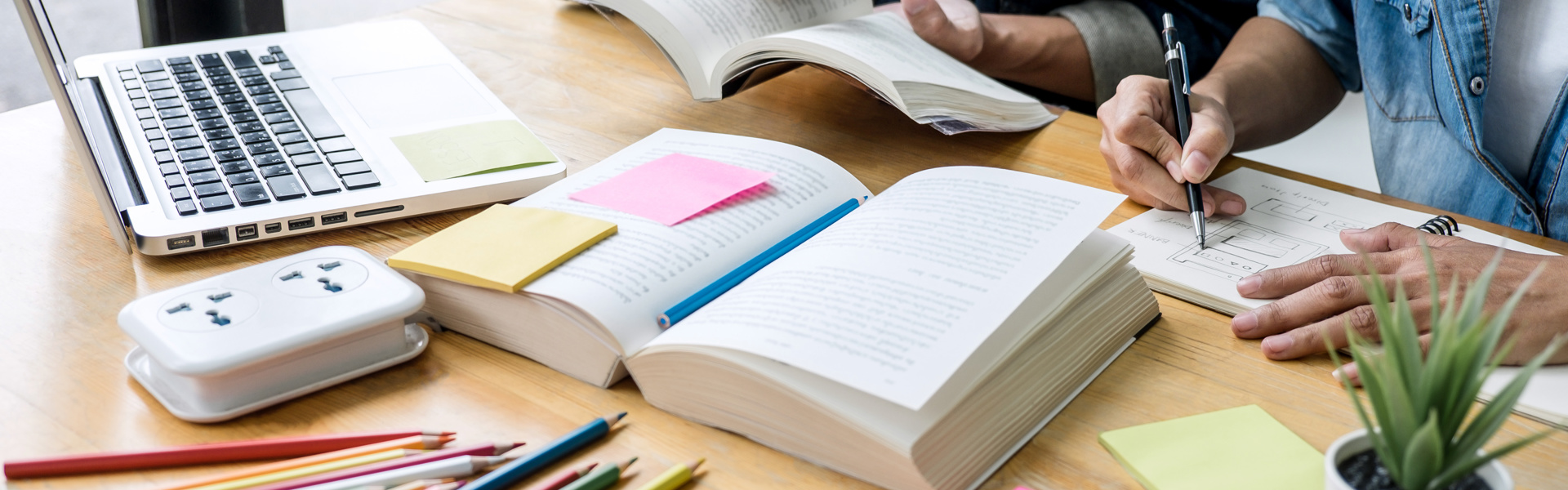 Desk with study equipment and stationary on it, two people sat at desk working together